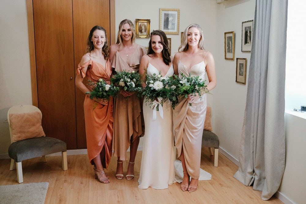Group of bridesmaids and bride with their bouquets of white and green flowers, and dresses in ochre, pastel, earth tones, matching the floor and room furniture
