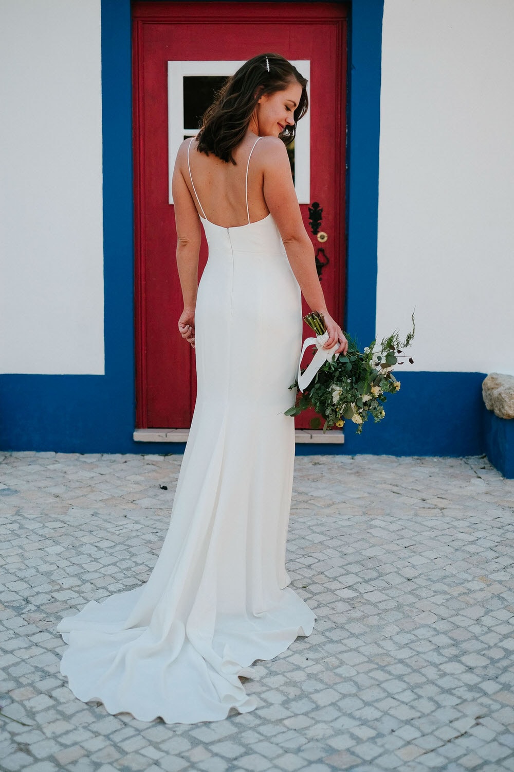 The bride without veil showcases the back of her dress and train while holding her bouquet in front of the door with a blue boat, a traditional Portuguese feature at Quinta da Bichinha
