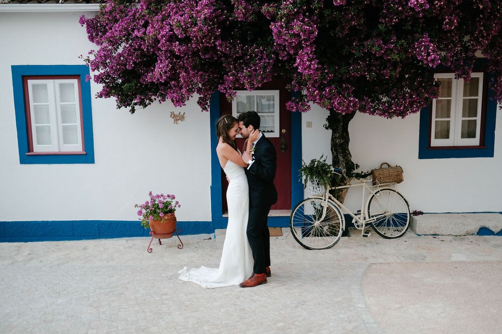 The groom and bride look at each other in front of a beautiful bougainvillea and a rustic bicycle in white, blue, and red colors of the quinta da bichinha with the typical Portuguese facade