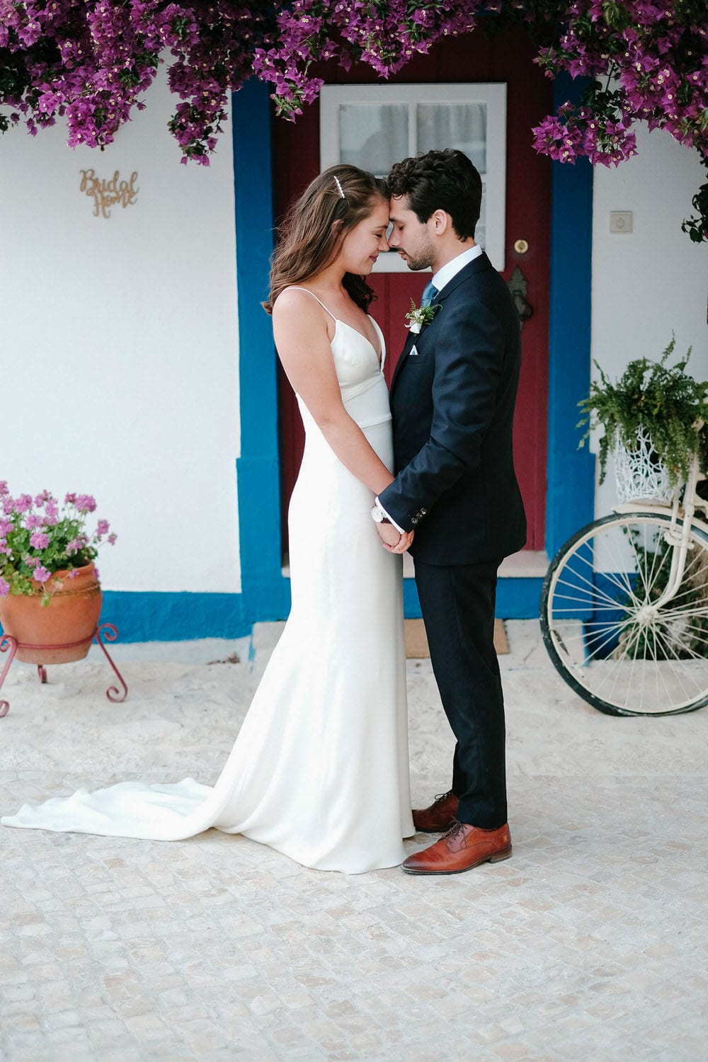 The bride and groom gaze at each other in front of a beautiful bougainvillea and a rustic bike in white, blue, and red at Quinta da Bichinha with a typical Portuguese facade