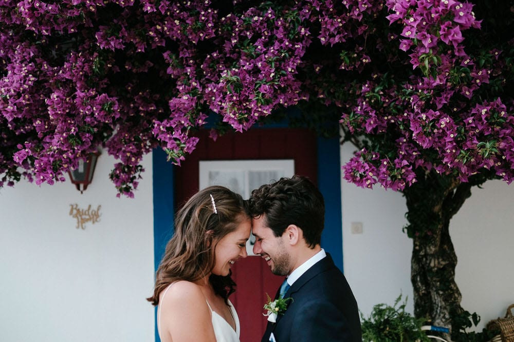 Quinta da Bichinha rustic destination wedding . The bride and groom gaze at each other in front of a beautiful bougainvillea and a rustic bike in white, blue, and red at Quinta da Bichinha with a typical Portuguese facade