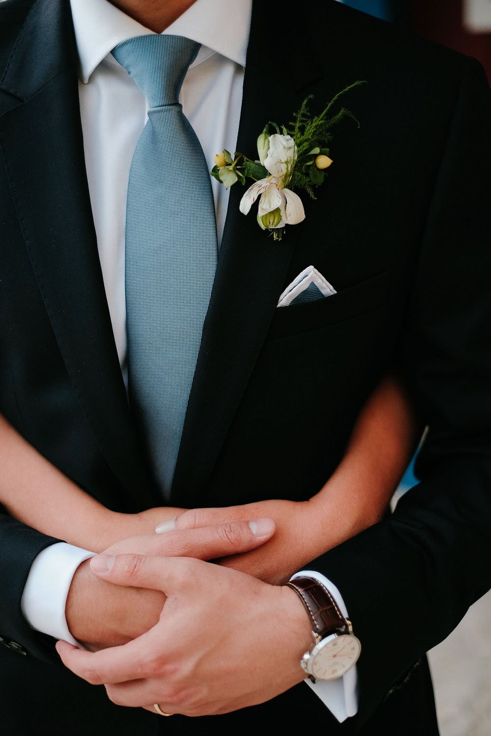 Close-up of the groom's lapel flower, along with his pocket square, tie, and the bride's intertwined hands with the groom's