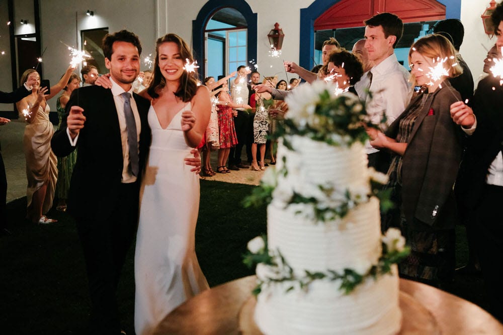 The couple looks at the wedding cake, surrounded by their guests with sparklers